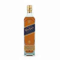 A single bottle of Johnnie Walker Blue Label whisky stands on a plain light gray background. The bottle is made of clear glass and shows the amber-brown liquid inside, with a dark blue label and gold details.
